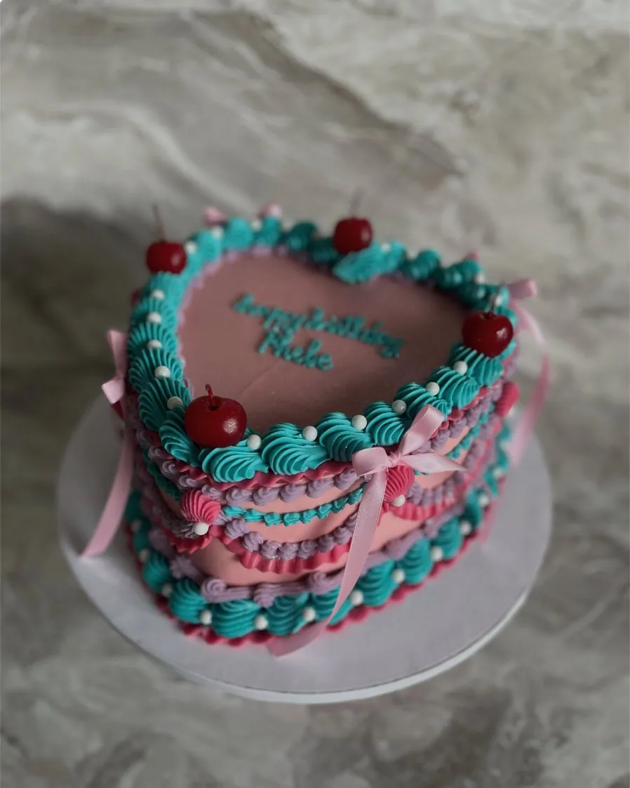 Pink birthday heart cake with teal shell piping, cherries and pink bows